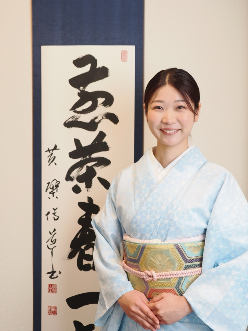 OCHARU's tea master, in kimono, beside a calligraphy scroll reading 'tea'